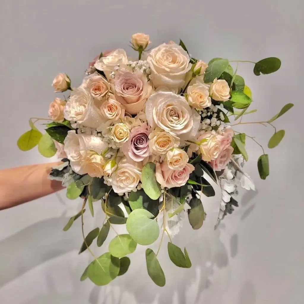 This image features a stunning bridal bouquet crafted by Casa D’Eramo, a vendor in the Wedding Decor category on Mariage Elegant, Canada. The bouquet is an elegant arrangement of blush and cream-colored roses, interspersed with delicate baby's breath and complemented by lush green eucalyptus leaves. The soft hues and varied textures create a classic and romantic feel, perfect for complementing a wedding ceremony setting.