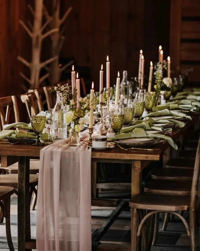 The image showcases a beautifully decorated wedding table by Casa D’Eramo, specializing in wedding decor. The table is elegantly set with a rustic wooden base, featuring tall, elegant taper candles and delicate floral arrangements in soft, natural tones. Green tinted glassware adds a touch of color, while neatly folded napkins complement the overall theme. A sheer, flowing table runner cascades along the center, enhancing the romantic and sophisticated atmosphere ideal for a wedding setting.