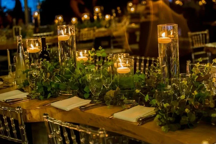 The image showcases an elegantly arranged wedding table set up by Casa D’Eramo, a vendor specializing in wedding decor. The table features tall, cylindrical glass vases with floating candles, creating a warm and inviting glow. Surrounding the vases are lush green foliage and small clusters of hydrangeas, adding a touch of natural beauty. Crystal-clear wine glasses and neatly folded white napkins are placed at each setting, complemented by the shimmering, transparent chairs. This stunning arrangement exemplifies Casa D’Eramo’s expertise in creating romantic and sophisticated decor for weddings.