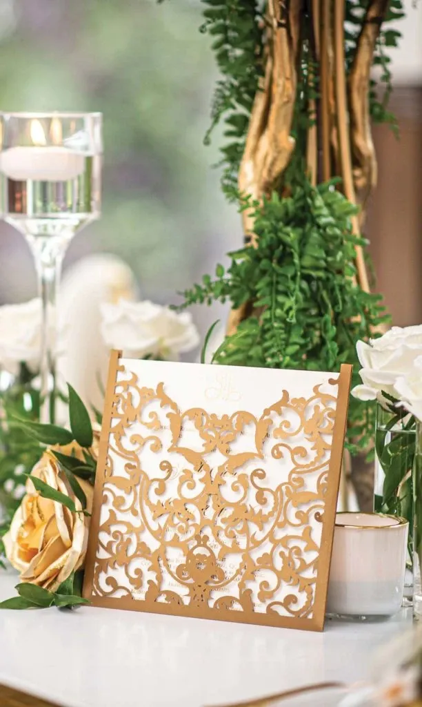 This image showcases an exquisite wedding decor arrangement by Casa D’Eramo in the wedding decor category. Featured prominently is an intricately designed gold filigree wedding invitation, resting elegantly on a table. The invitation's ornate pattern complements the surrounding decor, including white roses and lush greenery. A tall glass candle holder with a floating tealight adds a touch of sophistication, while trailing green vines and gold accents enhance the luxurious ambiance, ideal for a stylish and elegant wedding setting in Canada.