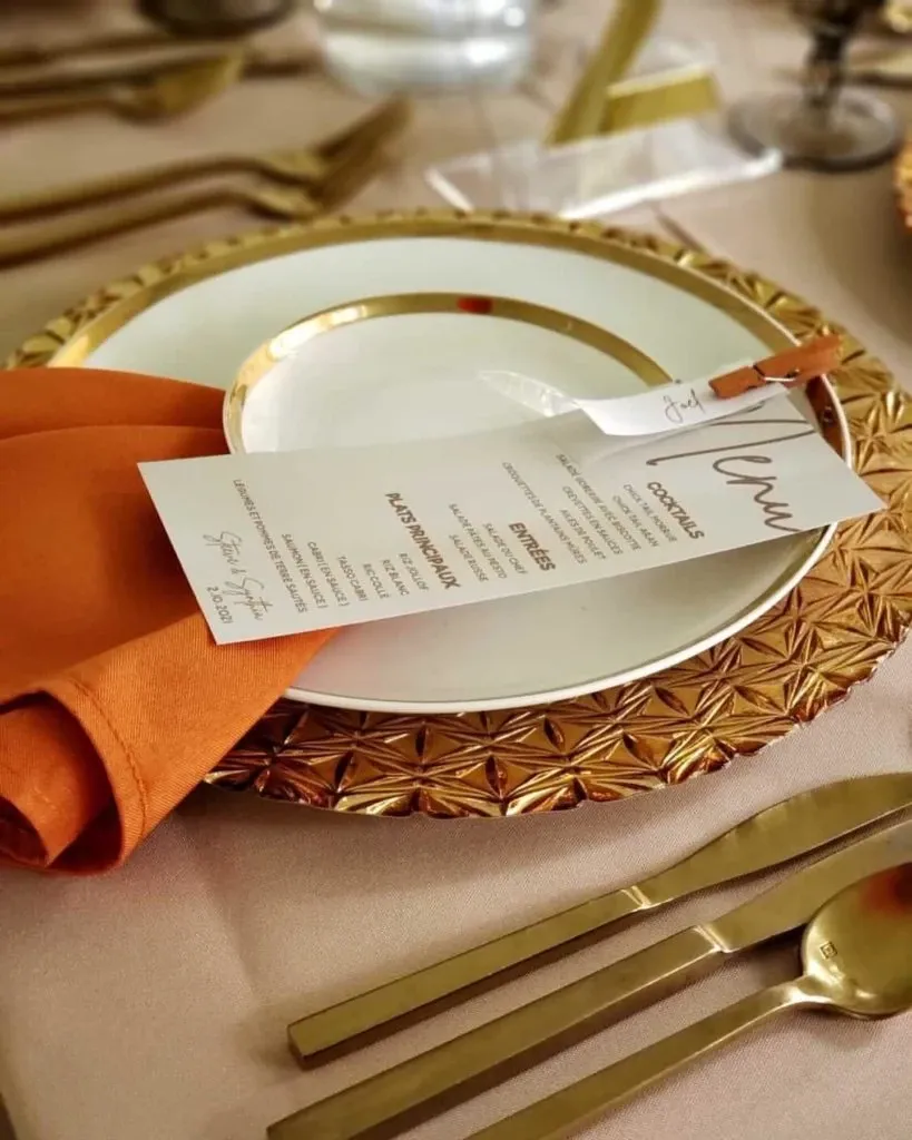 This image showcases an elegant wedding table setting by Casa D’Eramo in the wedding decor category. A beautifully designed place setting is featured, consisting of a white dinner plate with a gold rim, resting on an intricately patterned gold charger. An orange cloth napkin is elegantly folded beside the plate. A menu card with delicate calligraphy, listing various gourmet dishes and cocktails, is placed on the plate. Coordinated gold cutlery, including a knife and fork, is arranged on the beige tablecloth, enhancing the sophisticated and cohesive look.