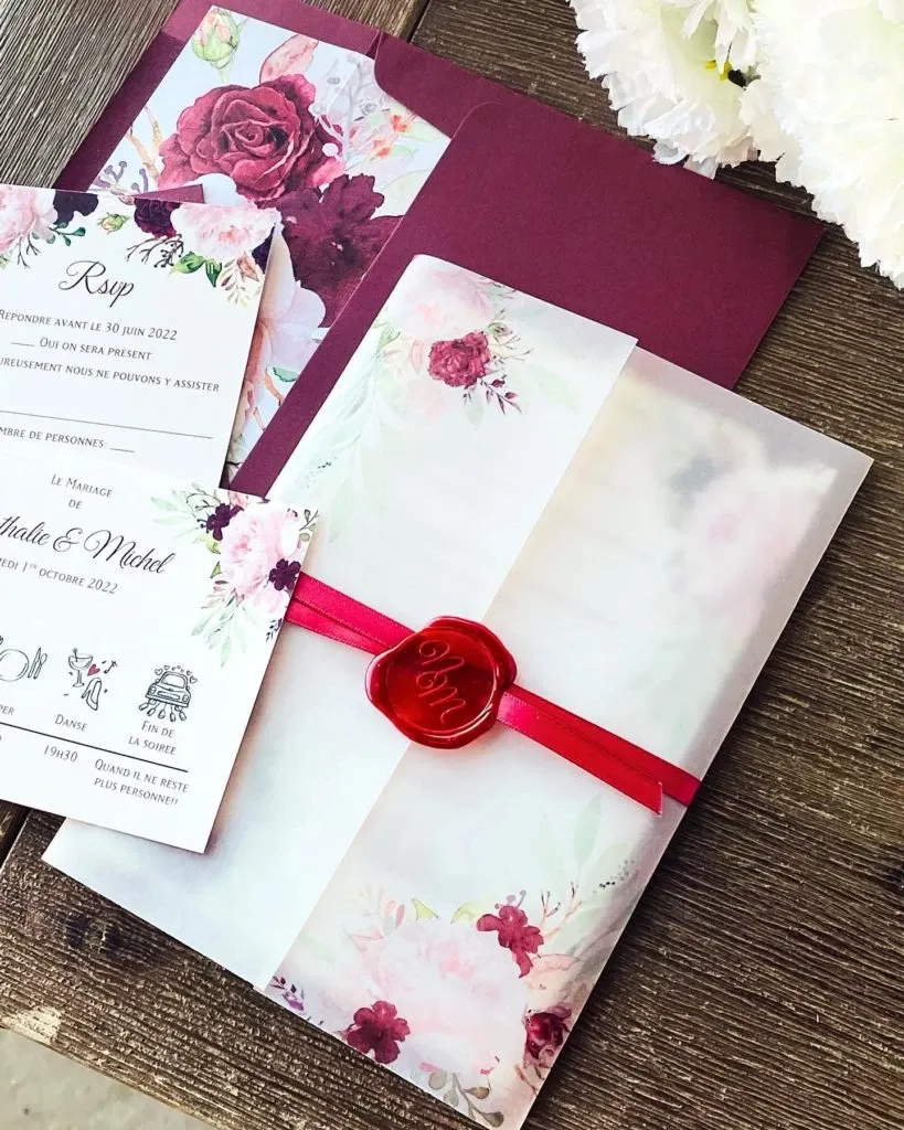 This image showcases a beautifully arranged wedding invitation set by Casa D’Eramo, under the Wedding Decor category. The centerpiece is an elegant invitation wrapped in translucent floral-patterned paper, secured with a vibrant red ribbon and a red wax seal imprinted with the word 'Love.' A rich burgundy envelope adds a touch of sophistication alongside a flat invitation card decorated with red and pink floral accents. The setting is complemented by white flowers in the background, emphasizing a romantic and refined aesthetic, suitable for a carefully planned wedding celebration by Mariage Elegant in Canada.