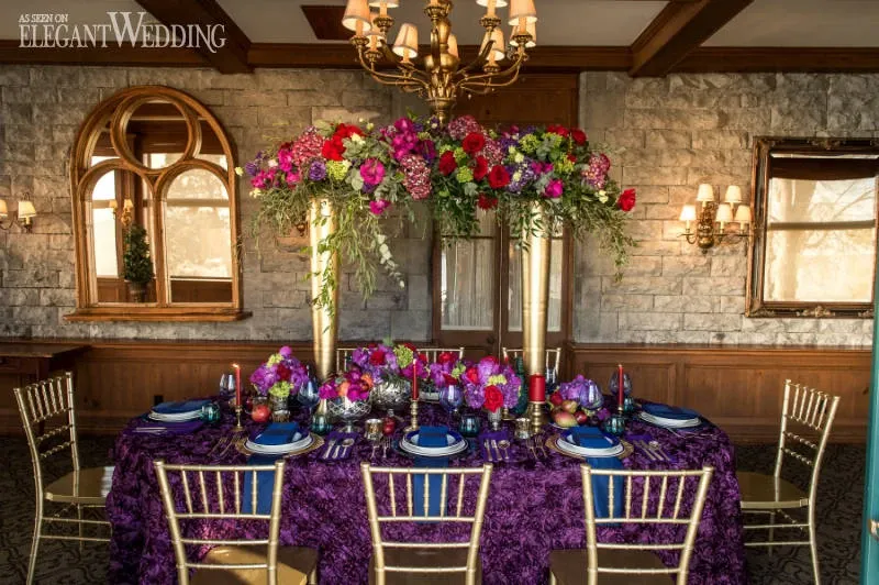 The image showcases a beautifully decorated wedding table by Casa D’Eramo, under the wedding decor category. The table is elegantly set with a deep purple tablecloth and gold chiavari chairs. Atop the table, there are vibrant floral arrangements featuring an array of pink, red, and purple flowers displayed in tall, gold stands, adding height and drama. The table settings include blue dinner plates, intricately designed glassware, and contrasting colorful napkins, creating a luxurious and opulent atmosphere. The surrounding decor includes rustic stone walls and elegant sconces, contributing to a sophisticated and romantic setting perfect for a wedding event.