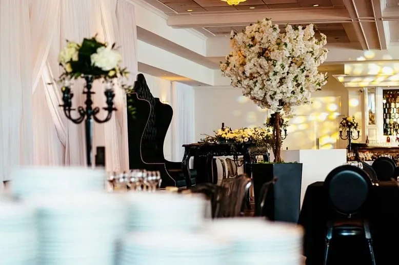 This image showcases a beautifully decorated wedding venue by Casa D’Eramo in the Wedding Decor category. The scene features an elegant arrangement with large, lush floral centerpieces dominated by white blossoms, enhancing the sophisticated ambiance. Black, ornate candelabras with white candles add a touch of classic elegance. In the background, luxurious black seating is complemented by soft, diffused lighting, creating a romantic atmosphere. A backdrop of sheer white drapery contributes to the overall refined aesthetic, making it perfect for an exquisite wedding celebration.