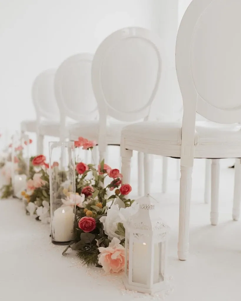 The image showcases elegant wedding decor by Casa D’Eramo in the wedding decor category. It features a row of classic white chairs with a curved back design, aligned neatly in a bright, softly lit setting. Alongside the chairs is a beautiful floral arrangement combining pink and white roses, lush greenery, and delicate petals accentuating the aisle. Interspersed among the flowers are tall glass candle holders and ornate white lanterns, creating a romantic and sophisticated atmosphere ideal for a wedding ceremony.