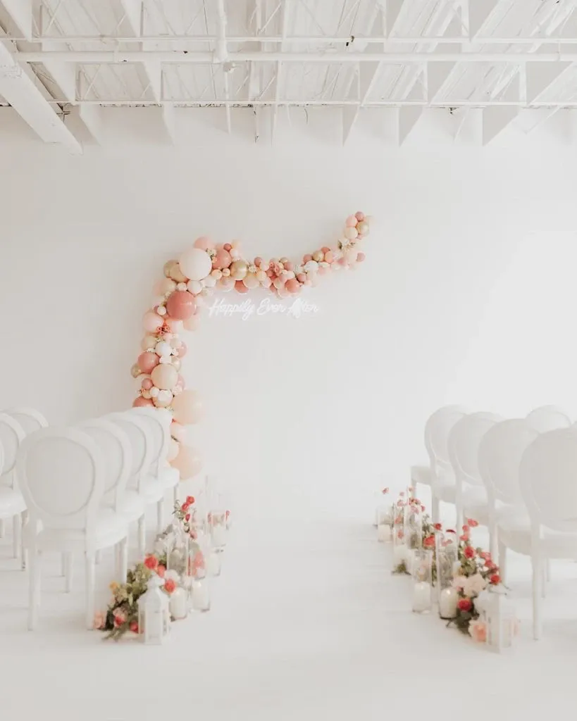 This image showcases an elegant wedding decor setup by Casa D’Eramo, featured in the wedding decor category on Mariage Elegant, Canada. The venue is adorned with a stunning balloon installation in soft pastel shades of peach and pink, elegantly cascading in an arch formation. Below the arch, a neon sign reads 'Happily Ever After,' adding a romantic touch to the setting. The aisle is beautifully lined with white chairs, and delicate flower arrangements and lanterns are placed thoughtfully along the path, creating an inviting and serene atmosphere for a wedding ceremony.