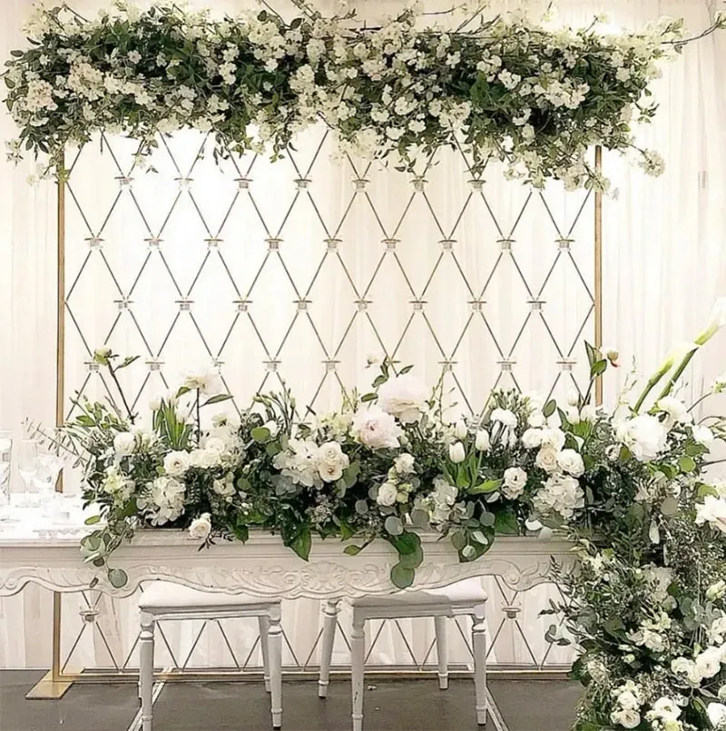 This elegant wedding decor image showcases a beautifully arranged setup by Decor and More Events in the wedding decor category. The focal point is an intricately designed sweetheart table adorned with lush white and green floral arrangements, featuring roses and other delicate blooms. Above the table, a matching floral arch elegantly frames the scene, creating a romantic atmosphere. The backdrop consists of a stylish white and gold patterned wall, adding a sophisticated touch to the overall decor. Perfect for elegant wedding celebrations, this display exemplifies fine craftsmanship and attention to detail.