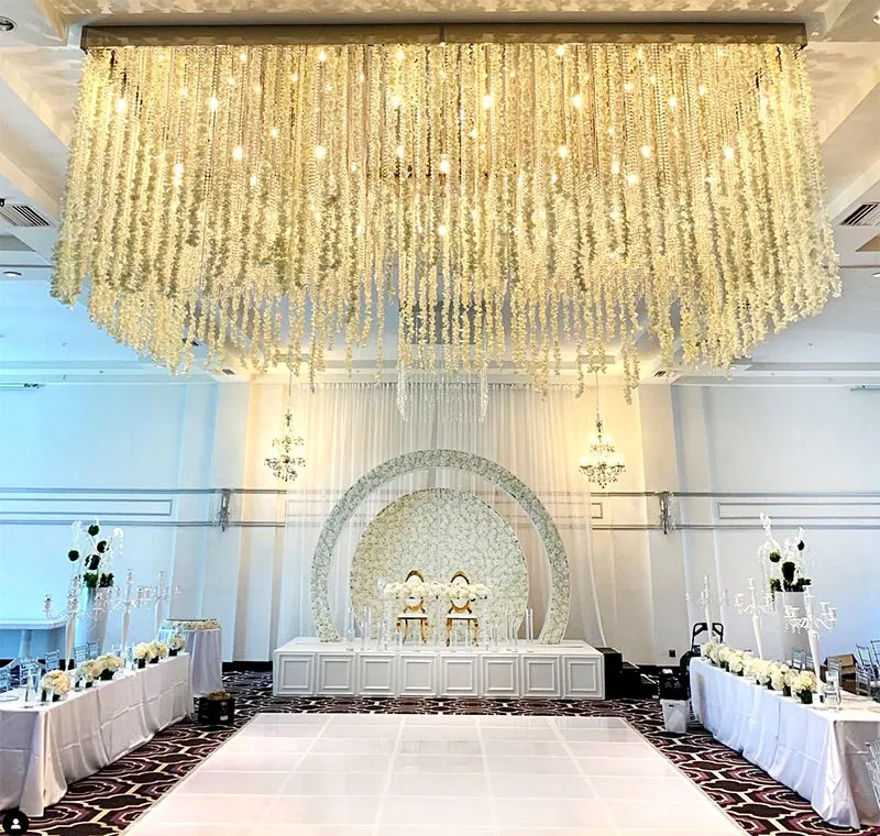 The image shows an elegantly decorated wedding venue by Decor and More Events under the wedding decor category. The setting features a grand chandelier canopy of cascading white flowers hanging from the ceiling, creating an ethereal atmosphere. A sophisticated backdrop with a circular floral arch is at the center, complementing the ivory and gold throne chairs positioned for the couple. Long tables are adorned with white floral arrangements and crystal candelabras, enhancing the luxurious theme. The venue showcases a pristine white dance floor, perfect for the couple’s first dance.