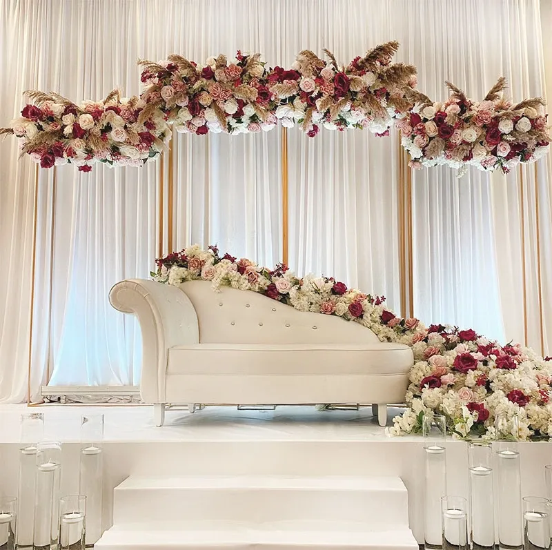 The image showcases an elegant wedding decor arrangement by Decor and More Events, categorized under Wedding Decor for Mariage Elegant in Canada. A luxurious white chaise lounge is prominently placed on a raised platform, surrounded by an abundance of floral arrangements. The flowers include a mix of white and deep pink roses, accented with pampas grass, creating a romantic and sophisticated ambiance. The background features soft, flowing white drapes that add a sense of elegance to the setting. Tall glass candle holders are arranged in front of the platform, contributing to the overall opulent and serene atmosphere of the wedding venue.