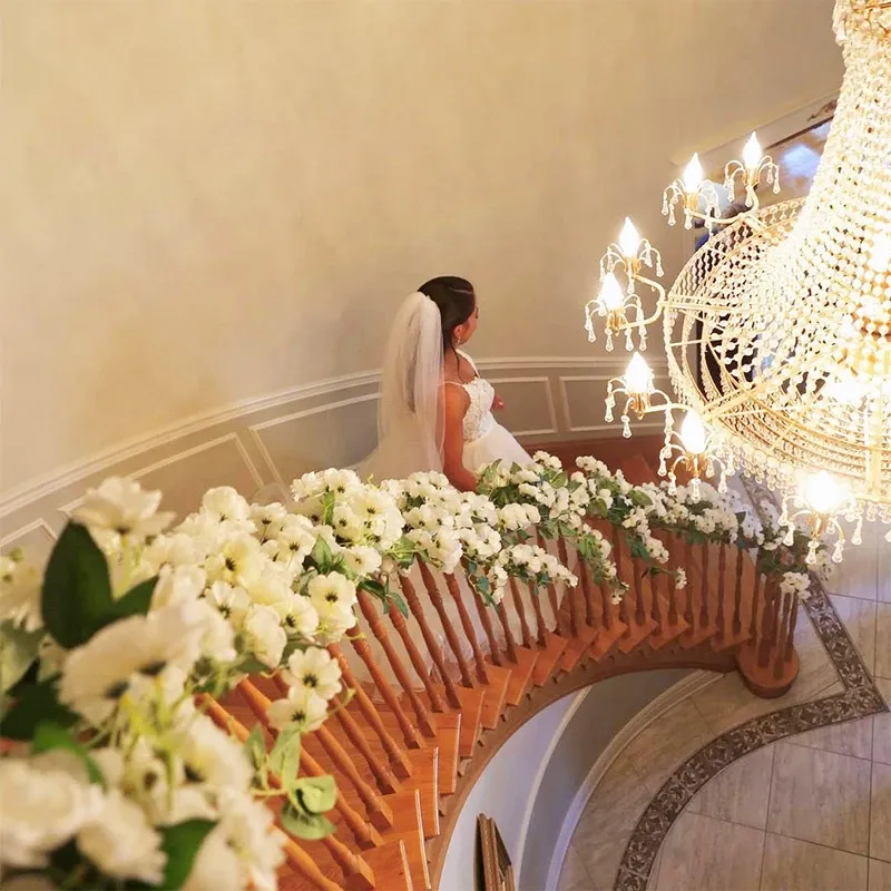 A bride in a white dress and veil descends a curved wooden staircase. The banister is elegantly adorned with lush arrangements of white flowers and greenery, typical of sophisticated wedding decor. Above, a large crystal chandelier casts a warm glow on the scene. This image showcases the exquisite wedding decor provided by Decor and More Events, highlighting their expertise in creating stunning and memorable settings for special occasions in the wedding decor category.