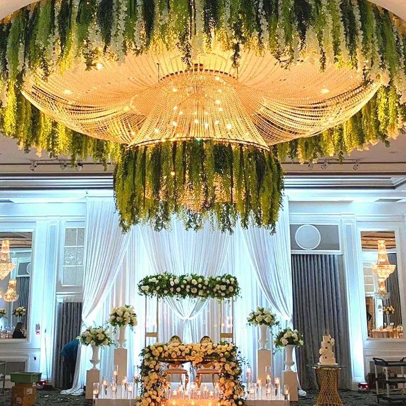 This image showcases an elegant wedding decor setup by Decor and More Events, categorized under Wedding Decor. The scene features a stunning, large chandelier adorned with cascading greenery, providing a lush and sophisticated ambiance. Beneath it, a backdrop of sheer white drapes creates a soft, elegant setting for the ceremony or reception. Arrangements of white roses and greenery are beautifully displayed on tall stands and across the front of a table that is surrounded by glowing candles. The overall design exudes a romantic and luxurious atmosphere fitting for an elegant wedding celebration.