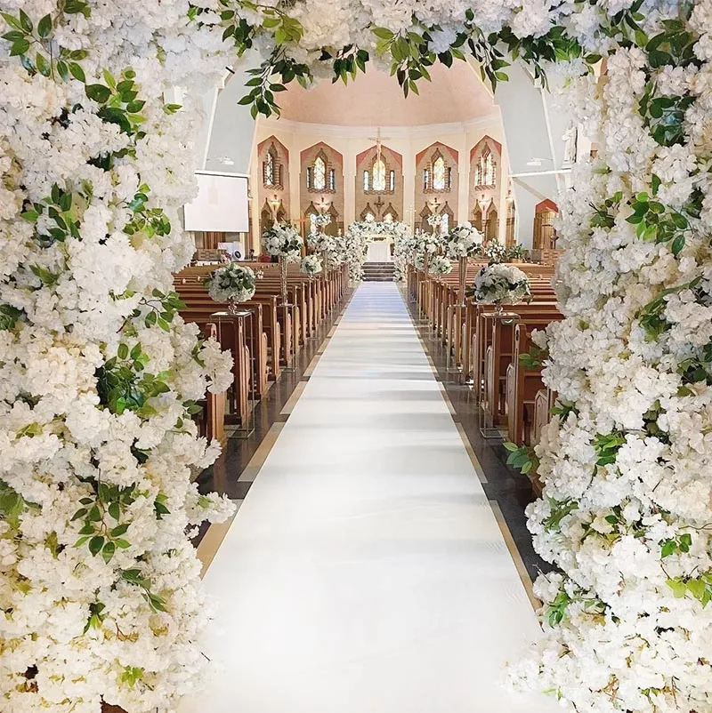 The image showcases a beautifully decorated wedding aisle created by Decor and More Events, categorized under Wedding Decor. The aisle is elegantly lined with lush white floral arrangements, forming a breathtaking archway that frames the entrance. The flowers extend along the length of the aisle, leading to a lavishly adorned altar at the front. The venue features wooden pews adorned with smaller matching floral bouquets, enhancing the sophisticated ambiance. The setting is completed with soft lighting and high ceilings, adding to the grandeur and elegance typical of a wedding event organized by Mariage Elegant in Canada.