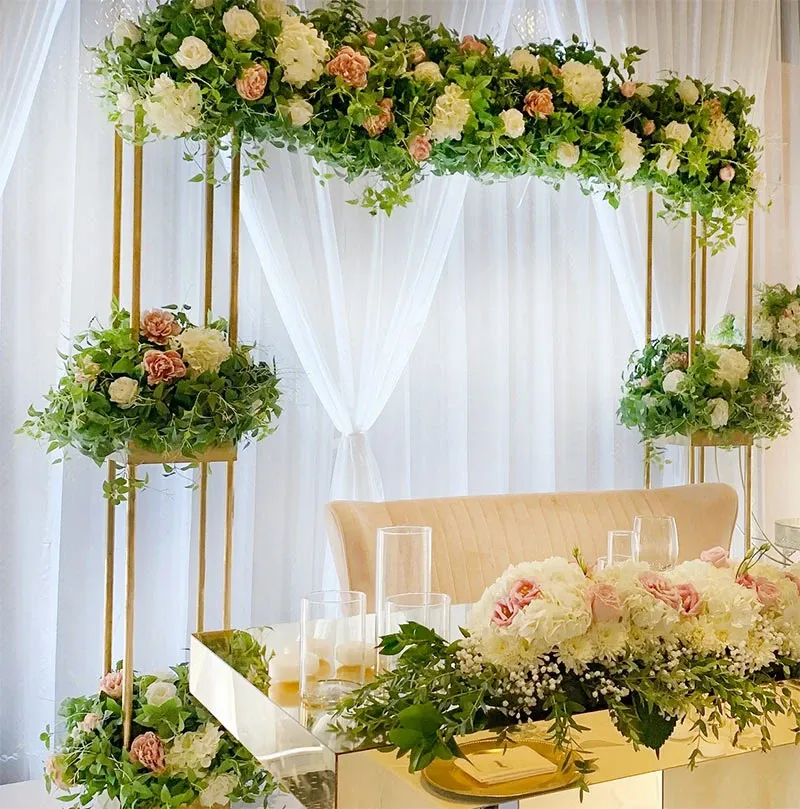 The image features a beautifully decorated wedding venue setup by Decor and More Events, categorized under Wedding Decor. It showcases an elegant floral arrangement with lush greenery and white and pink roses adorning a gold metallic structure. This structure serves as an arch over a luxurious table set against a backdrop of sheer white drapes. The table is embellished with matching floral centerpieces and clear glass candle holders, creating a sophisticated and romantic ambiance, ideal for a wedding celebration.