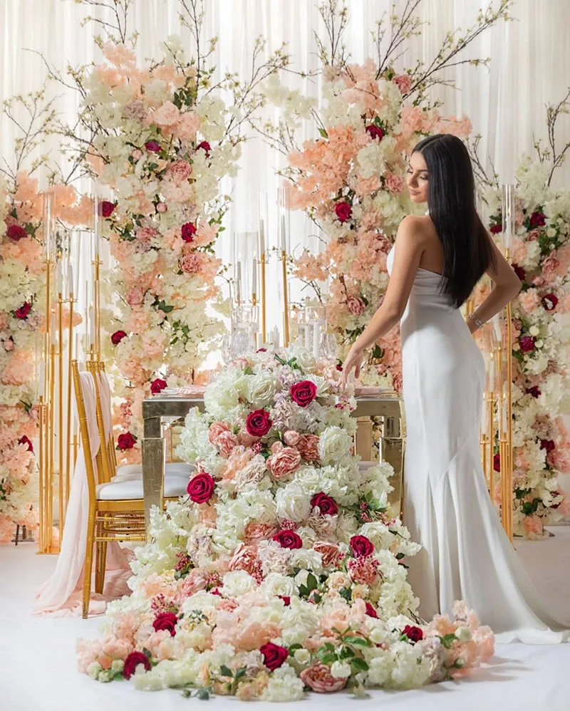 In this image showcasing the wedding decor by Decor and More Events for Mariage Elegant's vendor listing in Canada, a woman in an elegant white gown stands beside an opulently decorated table. The table is adorned with an abundant arrangement of flowers cascading to the floor, featuring a mix of blush pink, soft white, and deep red roses. Tall gold chairs add a touch of luxury, while the backdrop features tall floral displays with similar blooms, creating a romantic and sophisticated wedding ambiance. The overall setting exudes elegance and style, making it an ideal choice for couples seeking exquisite wedding decor.
