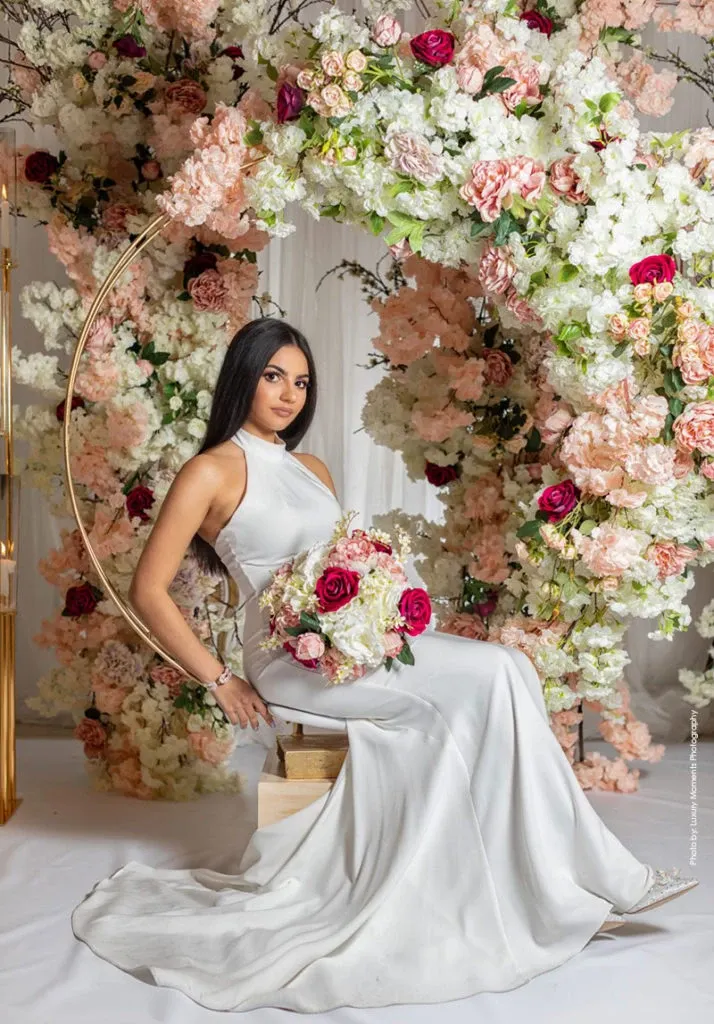 A bride in a sleek white gown is seated gracefully on a swing adorned with lush floral arrangements. The backdrop features elegant clusters of pink and white hydrangeas interspersed with vivid pink and red roses, creating a romantic and enchanting atmosphere. This exquisite setup showcases the artistry of Decor and More Events, a vendor listed under the Wedding Decor category on Mariage Elegant in Canada. The styling reflects a sophisticated and dreamy wedding theme, perfect for couples seeking elegance and charm.
