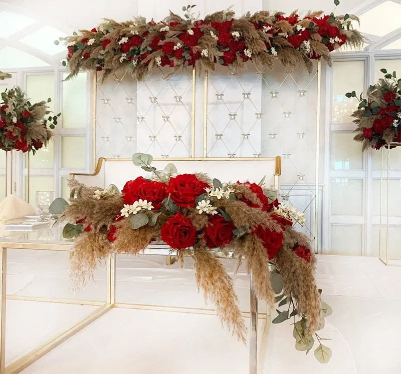 The image displays an elegant wedding decor setup by Decor and More Events, categorized under Wedding Decor for Mariage Elegant in Canada. It features a luxurious backdrop adorned with tufted white fabric panels and a lavish arrangement of red roses, pampas grass, and greenery cascading elegantly from the top. In the foreground, a matching display sits on a sleek, gold-accented table, combining red roses with delicate white flowers and softly textured pampas grass, creating a romantic and sophisticated ambiance perfect for weddings.