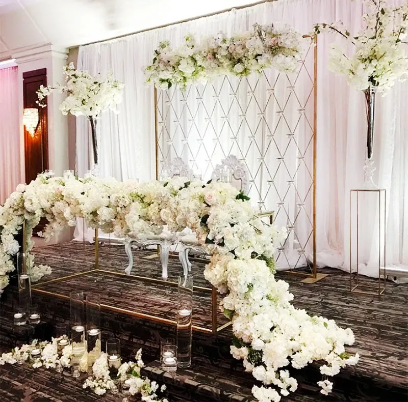 In the image, there is an elegant wedding decor setup by Decor and More Events, featured under the Wedding Decor category. A luxurious floral arrangement of white roses and hydrangeas cascades across a sophisticated metal arch structure adorned with delicate drapes. Tall floral arrangements flank each side, featuring similar white blooms that add height and elegance to the backdrop. In front of the arch, a stylish table and chairs are elegantly arranged, surrounded by glass vases with floating candles that enhance the romantic atmosphere. The overall design reflects a refined and classic approach, ideal for a high-end wedding setting by Mariage Elegant in Canada.