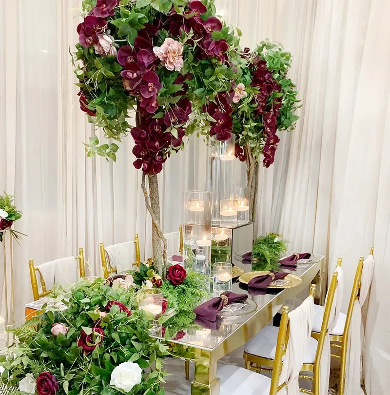 In this image from Mariage Elegant's vendor 'Decor and More Events,' the focus is on exquisite wedding decor. A luxurious table setup is showcased, featuring a long, reflective table adorned with elegant floral arrangements. Vivid burgundy and blush roses, along with rich green foliage, cascade from elevated structures above the table, creating a lush canopy. The table is set with gold-rimmed glasses and dark purple napkins, providing a sophisticated contrast. Tall, clear candle holders add a romantic glow, perfectly capturing the essence of an elegant wedding atmosphere.