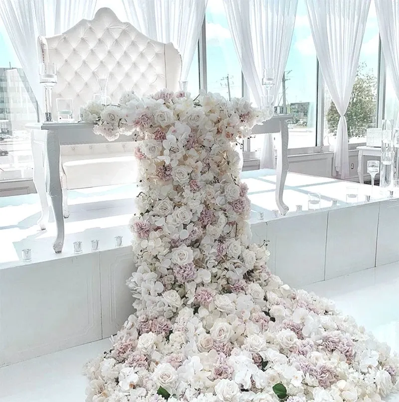 The image showcases an elegant wedding decor setup by Decor and More Events in the category of Wedding Decor. A luxurious white tufted high-back chair is positioned behind a white table, both set on an elevated platform. Cascading down the front of the table is a stunning arrangement of lush pink and white roses, creating a floral waterfall effect. The backdrop features sheer white drapes that allow natural light to filter through, adding to the overall romantic and sophisticated ambiance perfect for a wedding setting.