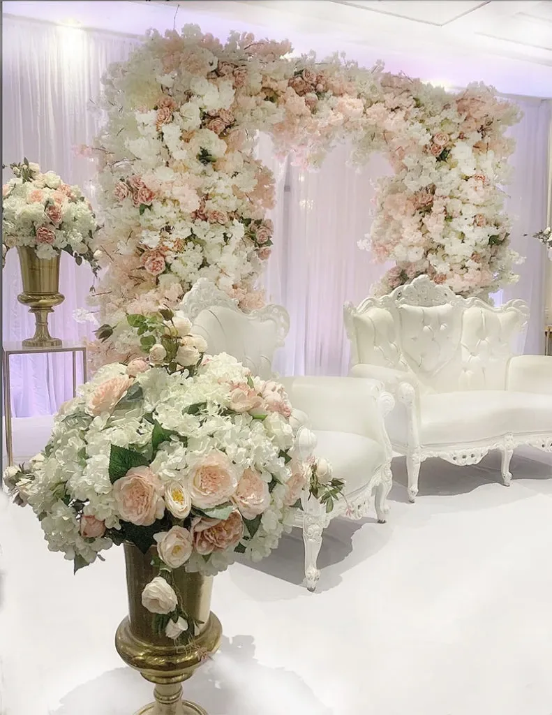The image features an elegant wedding decor setup by Decor and More Events, in the wedding decor category. A stunning floral archway, composed of white and blush pink flowers, serves as the focal point against a backdrop of sheer white curtains. In front of the arch, two ornately carved white chairs add a touch of sophistication. Gold vases brimming with matching floral arrangements are positioned on either side, enhancing the luxurious ambiance. This arrangement exemplifies high-end wedding decor suitable for elegant ceremonies and receptions.