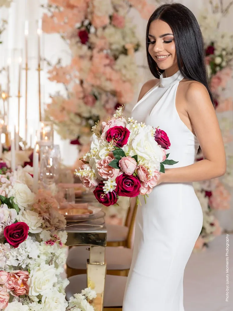 A bride holds a stunning bouquet featuring white, pink, and red roses, complemented by lush greenery. She stands beside an elegantly decorated table adorned with tall, gold candlesticks and a lavish floral arrangement in matching colors. The backdrop showcases an opulent display of flowers, contributing to the romantic and luxurious ambiance. This image exemplifies the exquisite work of Decor and More Events, highlighting their expertise in the Wedding Decor category for Mariage Elegant, Canada.