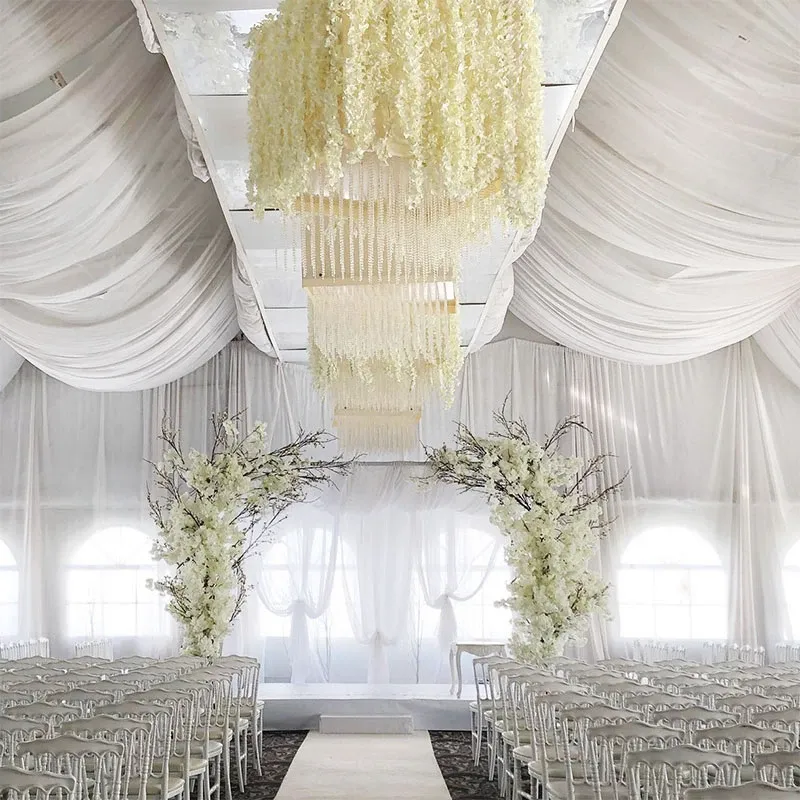 The image showcases an elegant wedding decor setup by Decor and More Events, falling under the wedding decor category on the Mariage Elegant site in Canada. The venue is beautifully adorned with white draped fabric cascading from the ceiling, creating a romantic and sophisticated atmosphere. A central aisle is flanked by rows of white chairs, leading to a stunning floral arch made of white blossoms. Above, a luxurious chandelier crafted from white flowers hangs, adding an opulent touch to the setting. Natural light filters through large windows, enhancing the airy and serene ambiance perfect for a memorable wedding ceremony.