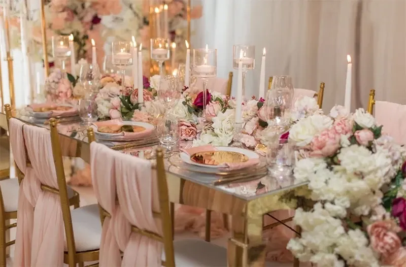The image showcases an elegantly arranged wedding decor setup by Decor and More Events, categorized under Wedding Decor. The scene features a luxurious table setting adorned with an array of lush white and pink floral arrangements, beautifully complementing the tall, elegant gold candle holders with lit tapered candles. Crystal glassware and gold-rimmed plates add a touch of sophistication, while soft pink draped chair covers create a harmonious and romantic atmosphere, perfect for a memorable wedding celebration. The overall aesthetic reflects an opulent and refined wedding event setup.