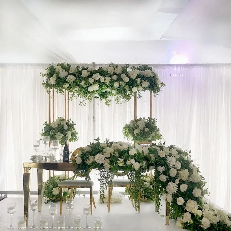This image showcases a beautifully arranged wedding decor setup by Decor and More Events, featured in the Wedding Decor category on Mariage Elegant, Canada. The scene includes an elegant arch draped with lush green foliage and white roses, creating a natural, romantic canopy over a stylish table setting. The table, accented with gold chairs, is adorned with cascading floral arrangements that mirror the arch above. Clear, cylindrical candle holders are placed strategically around the area, adding a touch of glamour and ambiance to the sophisticated design. The backdrop of sheer white curtains offers a serene and elegant atmosphere, perfectly suited for a wedding celebration.