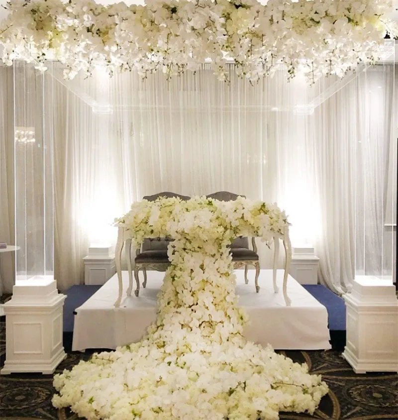 This image showcases an elegant wedding decor setup by Decor and More Events, featured in the wedding decor category. The scene includes a beautifully adorned table draped with cascading white floral arrangements, creating a luxurious and romantic ambiance. Above the table is a ceiling installation of white flowers, adding an ethereal touch to the decor. In the background, sheer white drapes softly frame the space, enhancing the overall elegance suitable for a sophisticated wedding celebration in Canada.