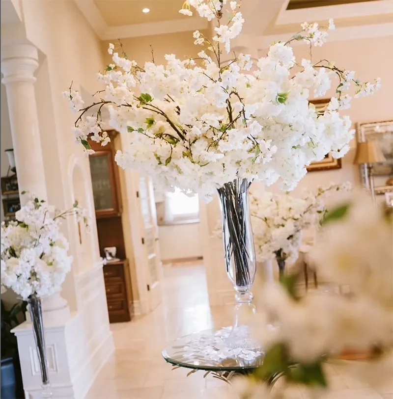 The image shows an elegant wedding decor setup by Decor and More Events, categorized under wedding decor. It features tall glass vases filled with abundant white blossoms, creating a lush and sophisticated atmosphere. The decor is arranged in a spacious, well-lit room with cream-colored walls and intricate architectural details, adding to the ambiance. This visually pleasing arrangement exemplifies luxury and attention to detail, reflecting the vendor's expertise in creating stunning wedding environments.