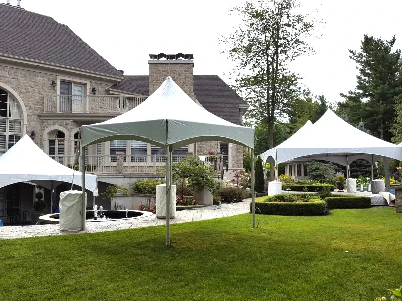 The image shows a beautifully landscaped garden area set up for a wedding, featuring elegant white tents provided by Chapiteau Montréal, a vendor in the Wedding Rentals category. The backdrop is a stately stone mansion with classic architectural details, framed by lush greenery and neatly manicured lawns. Several pristine tents are strategically placed on the grass, offering elegant cover for outdoor wedding events. The setting suggests a sophisticated venue, ideal for hosting a stylish and memorable wedding celebration.