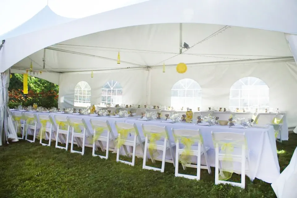 The image shows an elegantly arranged wedding reception setup under a large white tent provided by Chapiteau Montréal, specializing in wedding rentals. The interior features long tables covered in crisp white linens with neatly arranged white chairs embellished with soft yellow bows, capturing a serene and festive ambiance. Table settings include sparkling glassware and neatly arranged silverware. Soft lighting and yellow decorative accents contribute to the celebratory atmosphere, perfect for an outdoor wedding event in Canada. The tent is spacious, with arched windows allowing natural light to enhance the inviting decor.