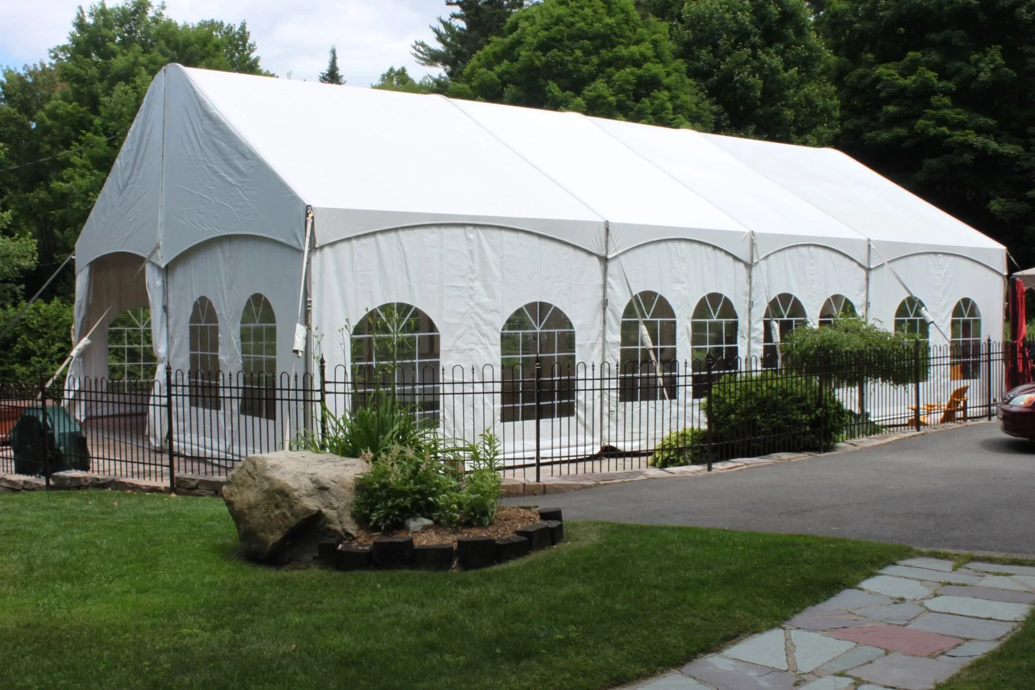 This image features a large, elegant white wedding tent provided by Chapiteau Montréal, a vendor in the Wedding Rentals category. The tent is set up outdoors on a neatly manicured lawn, surrounded by greenery and mature trees. It has white arched windows and is enclosed by a black wrought-iron fence. The spacious area suggests that it is ideal for a wedding reception or ceremony, offering a sophisticated and sheltered setting for guests. The scene conveys a tranquil and picturesque environment perfect for an elegant outdoor wedding event.