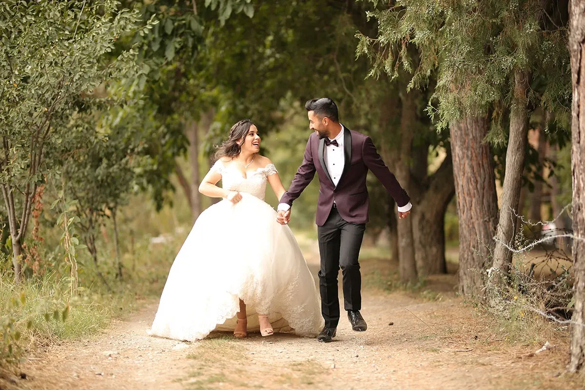 In this enchanting image, a joyful bride and groom walk hand in hand down a rustic, tree-lined path, evoking a sense of romance and celebration. The bride is wearing an elegant, flowing white gown, while the groom is dressed in a sophisticated dark suit with a bow tie. The lush greenery and serene natural setting create a perfect backdrop for their special day. This image showcases the wedding rentals offered by Chapiteau Montréal, highlighting their ability to enhance unforgettable wedding moments.