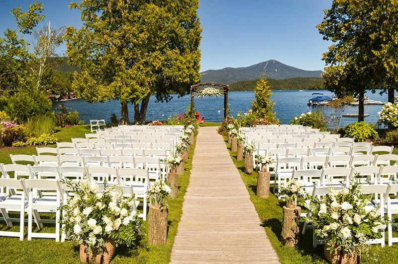 The image showcases an outdoor wedding setup provided by Chapiteau Montréal, categorized under Wedding Rentals. Set against a picturesque lakeside backdrop with mountains in the distance, the scene features rows of white chairs on either side of a wooden aisle. The aisle is adorned with rustic floral arrangements placed on tree stumps, creating a romantic and elegant atmosphere. Lush greenery and tall trees frame the setting, making it an ideal venue for a scenic outdoor ceremony.