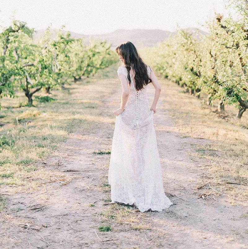 A bride in an elegant lace wedding gown walks gracefully down an aisle between blooming orchard trees. The soft sunlight casts a gentle glow on the scene, highlighting the intricate details of her dress. This serene outdoor setting suggests a romantic and natural backdrop, ideal for weddings. Chapiteau Montréal, a vendor specializing in wedding rentals, offers tents and other essentials to complement such picturesque venues.
