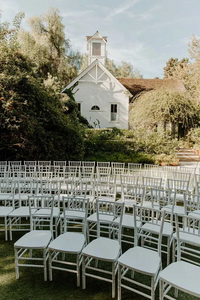This image showcases an elegant outdoor wedding setup provided by Chapiteau Montréal, categorized under Wedding Rentals. Rows of white chiavari chairs are neatly arranged on a lush green lawn, facing a charming white chapel that serves as a picturesque backdrop. The garden setting is surrounded by vibrant greenery and towering trees, adding a serene and romantic ambiance perfect for a wedding ceremony.