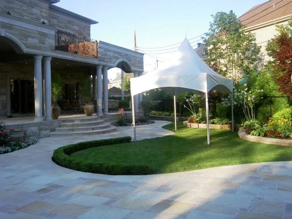 The image shows an elegant outdoor setting featuring a white canopy tent provided by Chapiteau Montréal, under the wedding rentals category. The tent is set up in a beautifully landscaped backyard with lush green grass and neatly trimmed hedges, adjacent to a stone building with classical architecture and tall columns. The stone patio area complements the sophisticated atmosphere, making it an ideal location for a wedding ceremony or reception. The surrounding colorful flowers and greenery enhance the romantic and inviting ambiance.