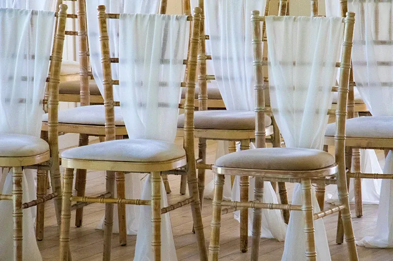 The image shows elegant wedding chairs provided by Chapiteau Montréal, a vendor in the Wedding Rentals category featured on Mariage Elegant, Canada. The chairs are wooden with a classic Chiavari design, featuring light cushion seats for added comfort. Each chair is draped with soft, flowing white fabric, adding a touch of sophistication and elegance, perfect for a wedding ceremony or reception setting. The chairs are arranged neatly in rows on a wooden floor, creating an inviting and polished atmosphere for guests.