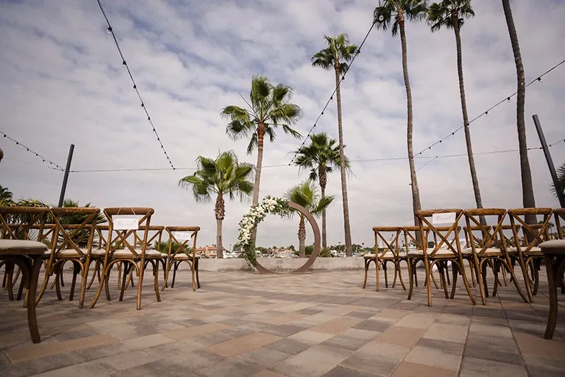 The image showcases a beautifully arranged outdoor wedding ceremony setup by Chapiteau Montréal, a wedding rentals vendor. It features an elegant arch adorned with lush greenery and white flowers, positioned at the center against a backdrop of tall palm trees and a cloudy sky. String lights are artistically draped overhead, adding a charming ambiance. Wooden chairs are neatly arranged in rows on a tiled patio, creating an inviting pathway towards the altar. This setup reflects a serene and romantic atmosphere, perfect for an outdoor wedding celebration.