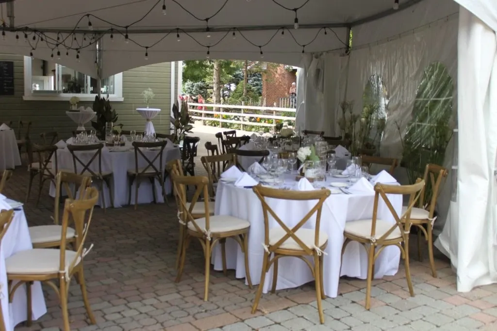 The image showcases a beautifully arranged outdoor wedding rental setup by Chapiteau Montréal, part of the wedding rentals category. Under an elegant tent, there are multiple round tables dressed in crisp white tablecloths, each adorned with chic table settings and glassware. Wooden cross-back chairs are neatly placed around the tables, providing a rustic yet sophisticated charm. Overhead, string lights create a warm, inviting ambiance, perfect for an intimate wedding reception. Lush greenery and decorative plants enhance the aesthetic, adding a touch of natural beauty to this well-coordinated setting provided by the Canadian vendor.