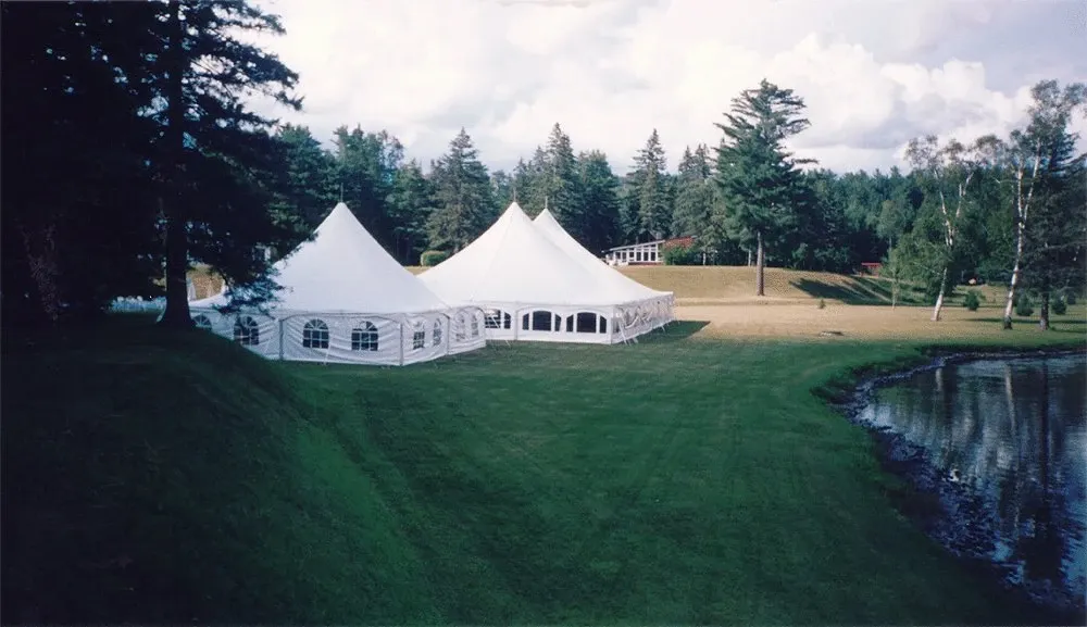The image features a large white wedding tent set up by Chapiteau Montréal, categorized under Wedding Rentals. The tent is situated on a lush green lawn near a serene pond, surrounded by tall trees, creating a picturesque outdoor wedding setting. The structure has elegant peaks and arched windows, providing a spacious and sheltered area for wedding receptions or ceremonies. Fluffy clouds are visible in the blue sky, adding to the tranquil and romantic atmosphere ideal for a memorable wedding event.