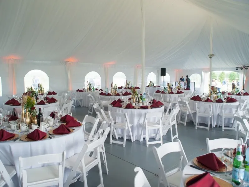 The image shows an elegantly arranged wedding reception setup provided by Chapiteau Montréal, a vendor in the Wedding Rentals category. Inside a large, white tent, round tables are neatly adorned with deep burgundy napkins and elegant centerpieces featuring candles and floral arrangements. The luminous, white drapery and soft ambient lighting create a sophisticated and inviting atmosphere. White folding chairs are neatly arranged around the tables, enhancing the overall elegance suitable for a formal wedding event.