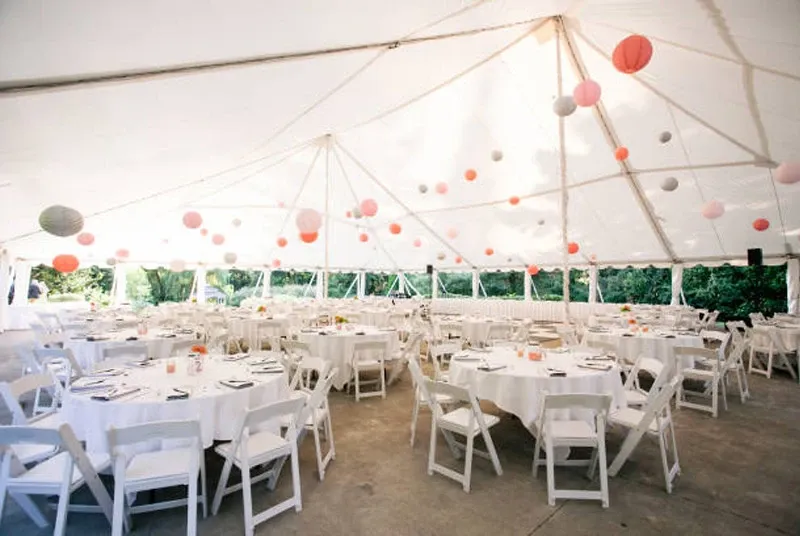 The image displays a beautifully arranged outdoor wedding tent set up by Chapiteau Montréal, featured in the Wedding Rentals category on Mariage Elegant, Canada. Inside the spacious tent, round tables are elegantly decorated with white tablecloths and neatly arranged white chairs. Each table is set with polished glassware and arranged plates, suggesting a sophisticated wedding reception setup. The ceiling of the tent is adorned with colorful paper lanterns in shades of pink, red, and gray, adding a festive touch to the ambiance. The surrounding greenery is visible through the sides of the tent, offering a picturesque and serene backdrop for the event.