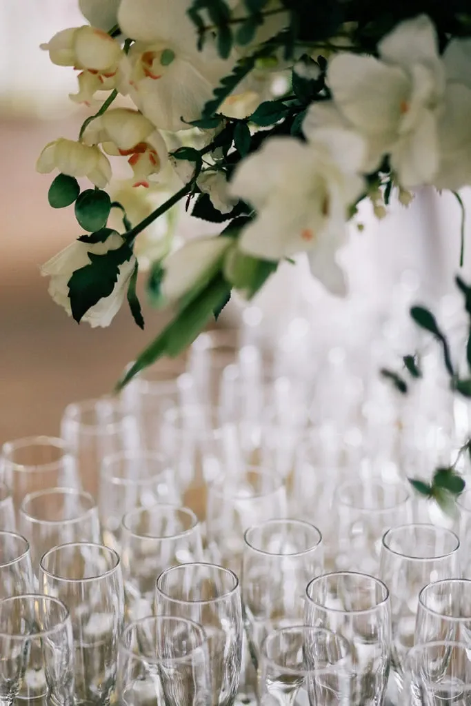 The image features an elegant arrangement of clear champagne glasses neatly aligned on a table, ready for a wedding celebration. Above them, a lush floral arrangement showcases delicate white orchids with soft green leaves, adding a touch of elegance. This setup is part of the wedding rentals offered by Chapiteau Montréal, showcasing their attention to detail and ability to enhance the ambiance of a wedding venue. The overall presentation conveys sophistication, perfect for a stylish and memorable event.