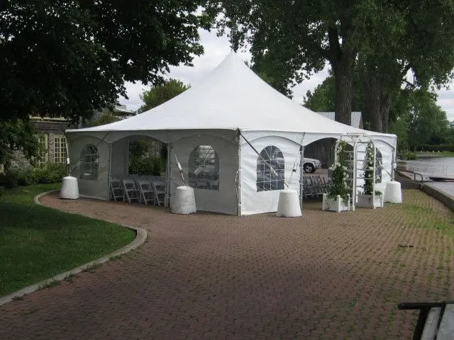 This image features a large white wedding tent set up by Chapiteau Montréal, categorized under Wedding Rentals. The tent is elegantly structured with a peaked roof and clear plastic windows, allowing natural light to illuminate the interior. It is situated on a spacious, paved area surrounded by lush greenery and mature trees, creating a serene outdoor setting ideal for weddings. Within the tent, rows of white chairs are neatly arranged, indicating a readiness for hosting guests. The scenic backdrop of a waterfront further enhances the picturesque wedding venue atmosphere.