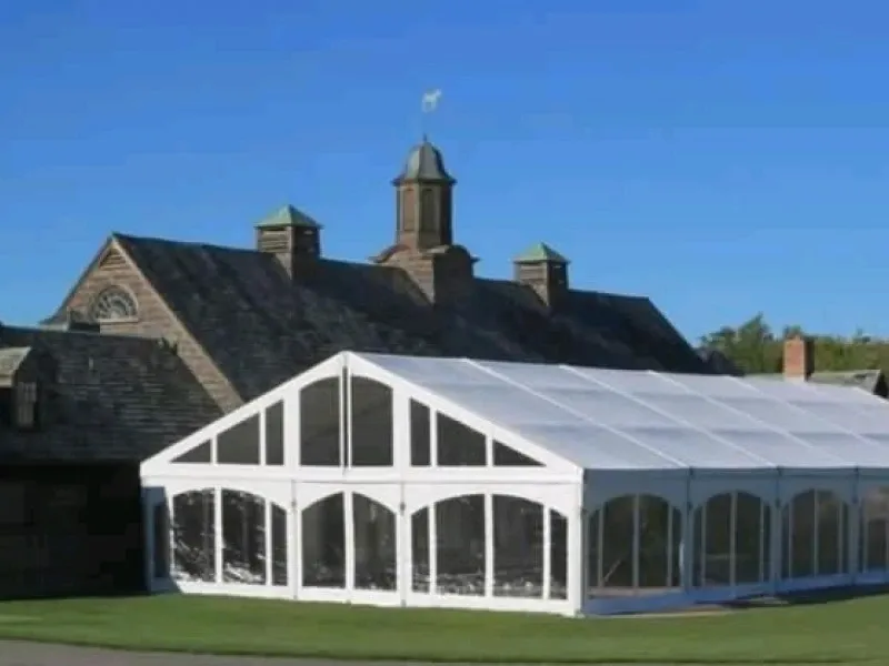 The image shows a large white wedding tent set up by Chapiteau Montréal, categorized under Wedding Rentals. The tent is pitched on a lush green lawn in front of a picturesque stone building with a shingled roof and multiple skylights. The structure of the tent features transparent sides and a gabled roof, allowing natural light to filter through. The setting is serene and elegant, ideal for a wedding ceremony or reception, capturing the essence of outdoor luxury wedding events in Canada.