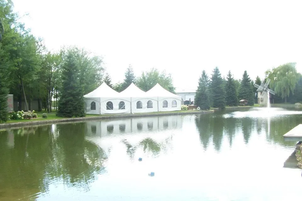 The image showcases an elegant white tent setup by Chapiteau Montréal, a prominent vendor in the Wedding Rentals category. Positioned by a serene lake, the large tent features arched window panels that allow natural light to filter through. Surrounded by lush greenery and tall trees, the setting creates a picturesque and tranquil atmosphere ideal for a wedding celebration. The reflection of the tent and trees in the water adds to the scenic beauty, making it a perfect choice for outdoor events.