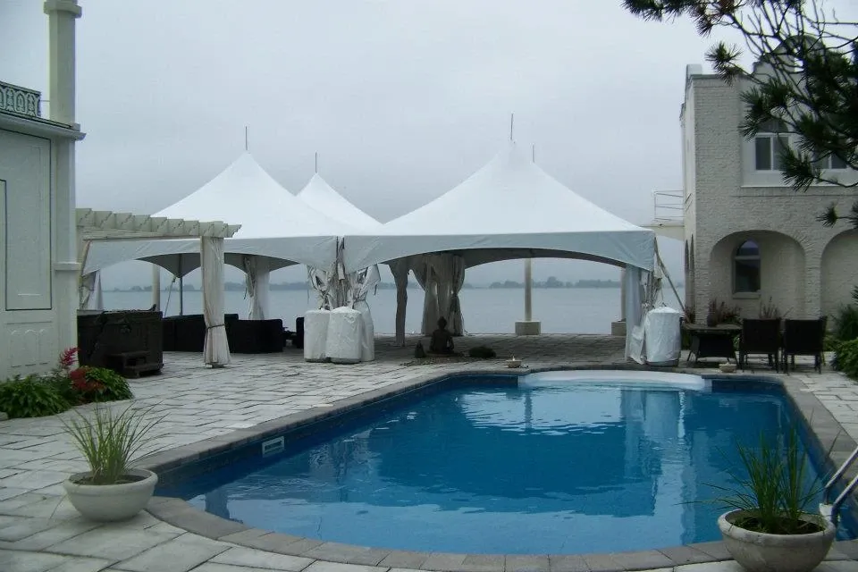 The image showcases an elegant outdoor setting featuring multiple white tents provided by Chapiteau Montréal, categorized under Wedding Rentals for Mariage Elegant, Canada. These tents are situated poolside on a spacious stone patio, adjacent to a large body of water, offering a picturesque and serene backdrop ideal for a wedding celebration. The area is accented with potted plants and sophisticated patio furniture, enhancing the upscale ambiance of the venue.