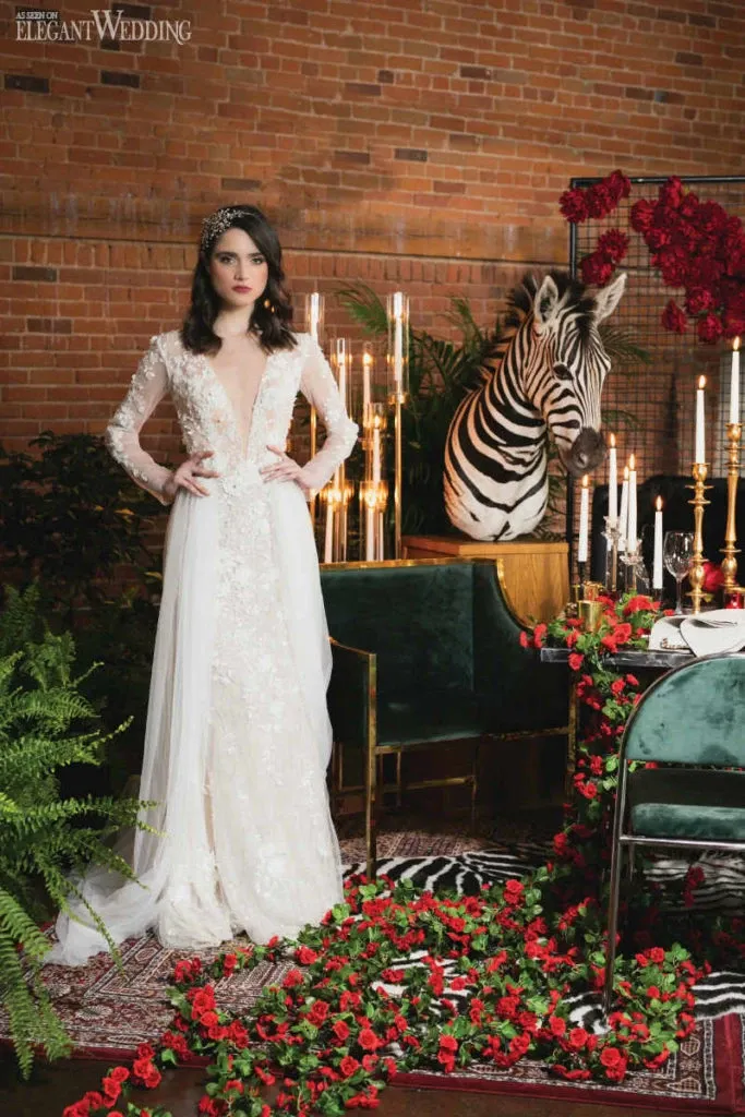In this image from Studio Caroline Théoret, featured in the Wedding Beauty category for Mariage Elegant, Canada, a bride stands elegantly in a long-sleeved, lace wedding gown with a deep V-neckline, amidst an opulent setting. Her dark hair is styled with a sparkling headpiece. To her left, a zebra adds an unexpected, whimsical touch, standing near a table adorned with tall, white candles and lavish floral arrangements dominated by red roses. The brick wall backdrop and lush greenery enhance the romantic and luxurious ambiance, making it a unique setting for wedding photography and beauty contemplation.